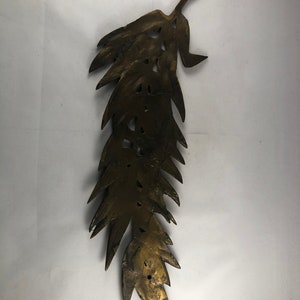 May include: A gold-colored metal leaf-shaped wall decor with a detailed design. The leaf is long and narrow with a pointed tip. It has a rustic, aged look.