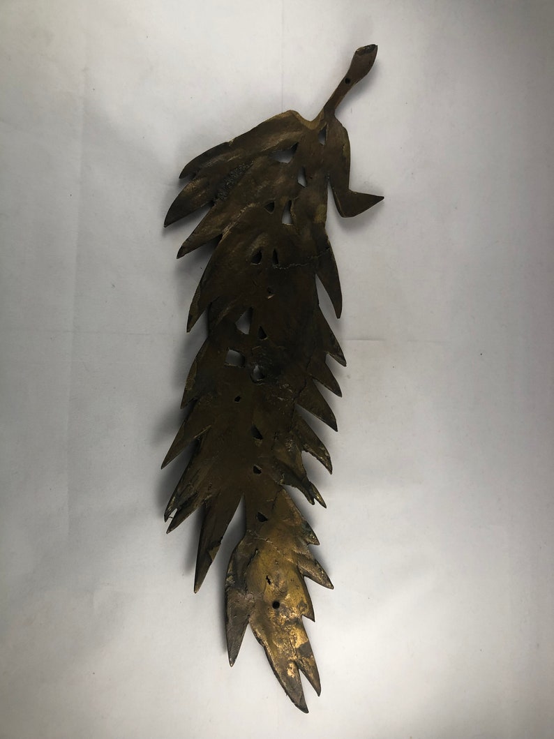 May include: A gold-colored metal leaf-shaped wall decor with a detailed design. The leaf is long and narrow with a pointed tip. It has a rustic, aged look.