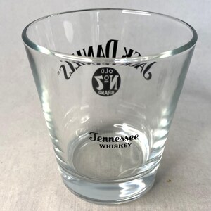 May include: Clear glass tumbler with the words "Jack Daniel's" and "Tennessee Whiskey" printed in black. The glass has a slightly flared shape and a thick base.