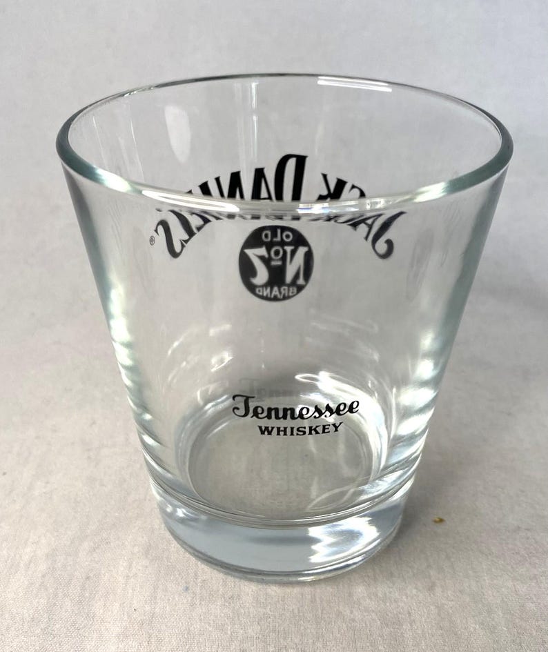 May include: Clear glass tumbler with the words "Jack Daniel's" and "Tennessee Whiskey" printed in black. The glass has a slightly flared shape and a thick base.