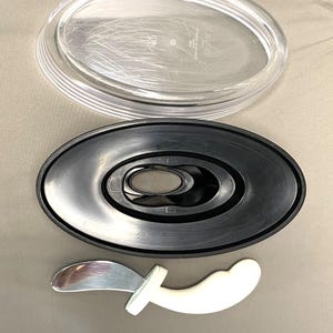 May include: A clear plastic oval lid and a black oval base with a central opening are displayed. Below, a small, white, curved handle with a silver blade is visible. These items are likely parts of a serving dish or kitchen utensil.