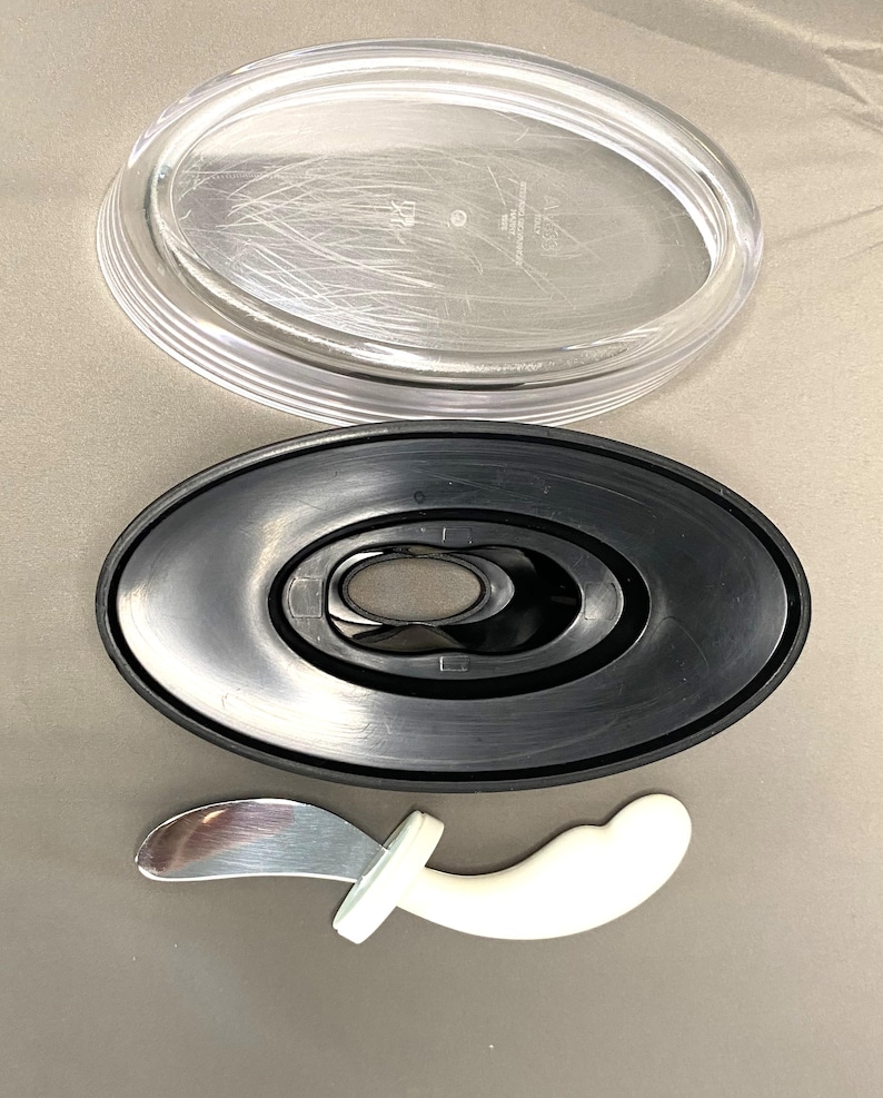 May include: A clear plastic oval lid and a black oval base with a central opening are displayed. Below, a small, white, curved handle with a silver blade is visible. These items are likely parts of a serving dish or kitchen utensil.