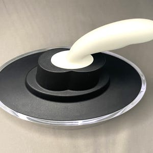 May include: A bottle stopper with a black base and a clear, rounded edge. A white, curved handle extends from the centre. The stopper is designed to fit on a bottle.