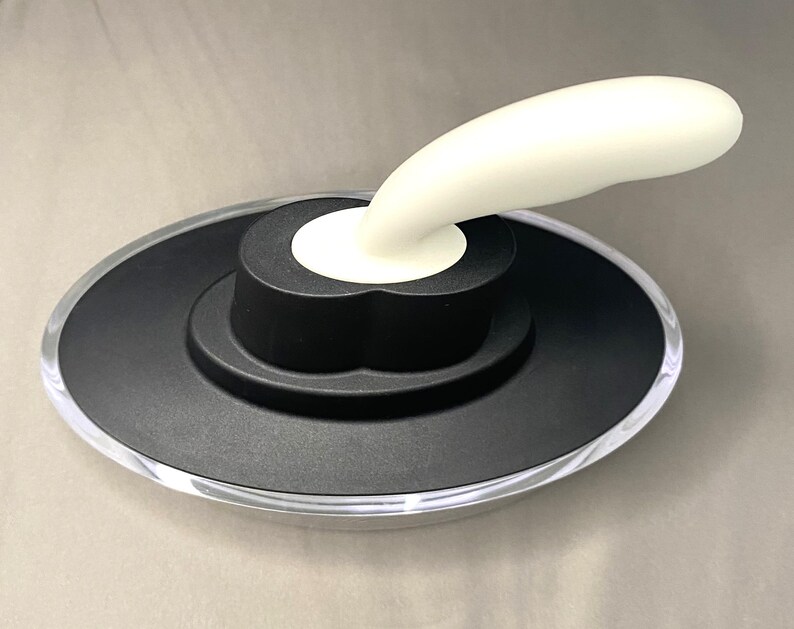 May include: A bottle stopper with a black base and a clear, rounded edge. A white, curved handle extends from the centre. The stopper is designed to fit on a bottle.