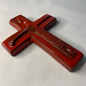 May include: A glossy, red ceramic cross with a detailed design. The cross has a rectangular shape with rounded edges and a dark brown accent. The religious symbol is displayed on a white background.
