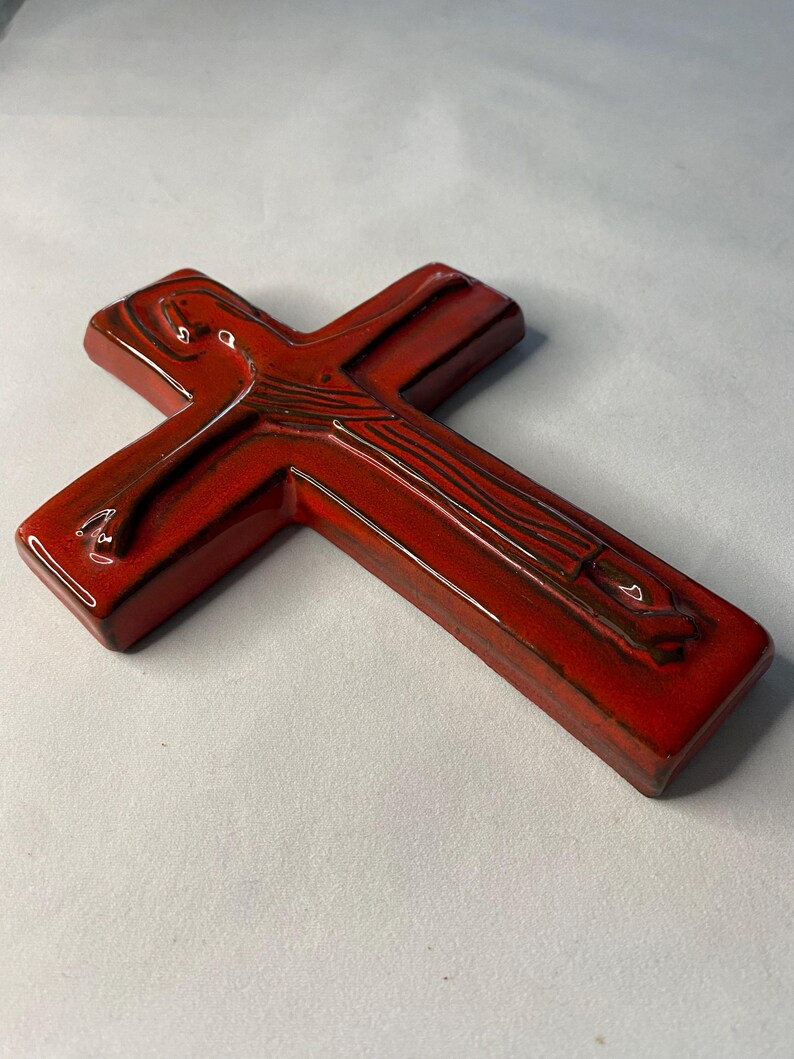 May include: A glossy, red ceramic cross with a detailed design. The cross has a rectangular shape with rounded edges and a dark brown accent. The religious symbol is displayed on a white background.