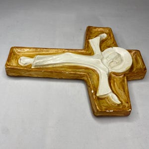 May include: A religious wall cross with a figure of Jesus. The cross is a warm brown color with a white figure. The figure is lying down with a halo around the head. The cross is made of a solid material.