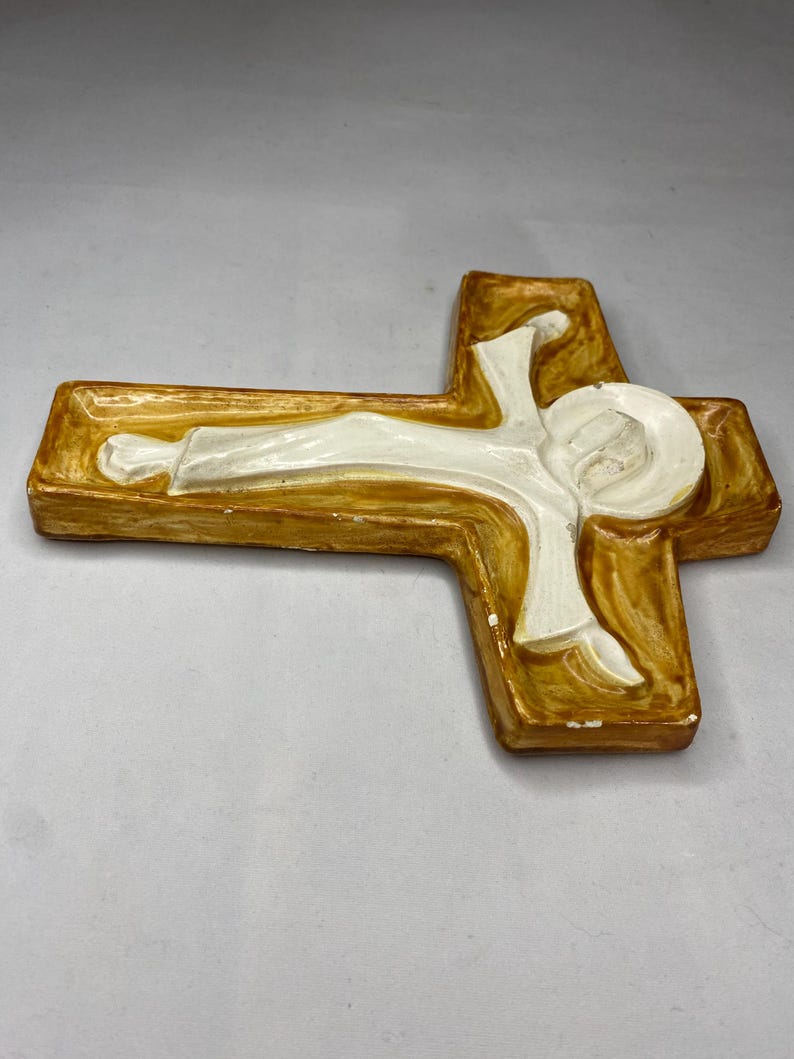 May include: A religious wall cross with a figure of Jesus. The cross is a warm brown color with a white figure. The figure is lying down with a halo around the head. The cross is made of a solid material.