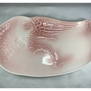 May include: A pink ceramic serving dish shaped like a fish with wings. The dish has a textured surface and is decorated with a raised design.