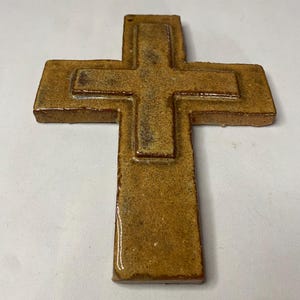 May include: A brown ceramic cross with a raised design. The cross is made of a textured material and has a glossy finish.