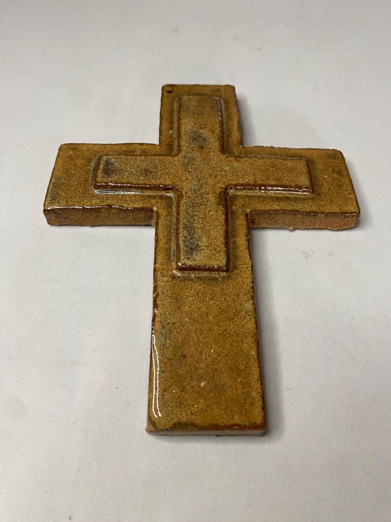 May include: A brown ceramic cross with a raised design. The cross is made of a textured material and has a glossy finish.