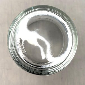 May include: A clear glass jar, seen from above, with a wide opening and a slightly curved interior. The jar's rim is thick and has a series of concentric rings. The glass reflects light, creating bright highlights and shadows.
