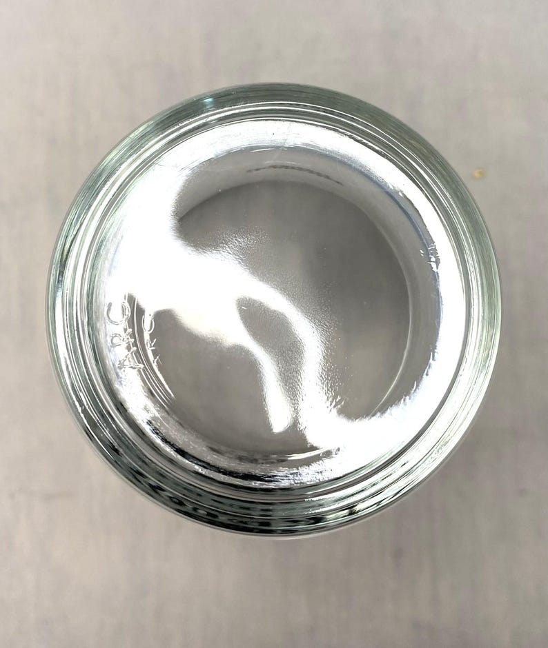 May include: A clear glass jar, seen from above, with a wide opening and a slightly curved interior. The jar's rim is thick and has a series of concentric rings. The glass reflects light, creating bright highlights and shadows.