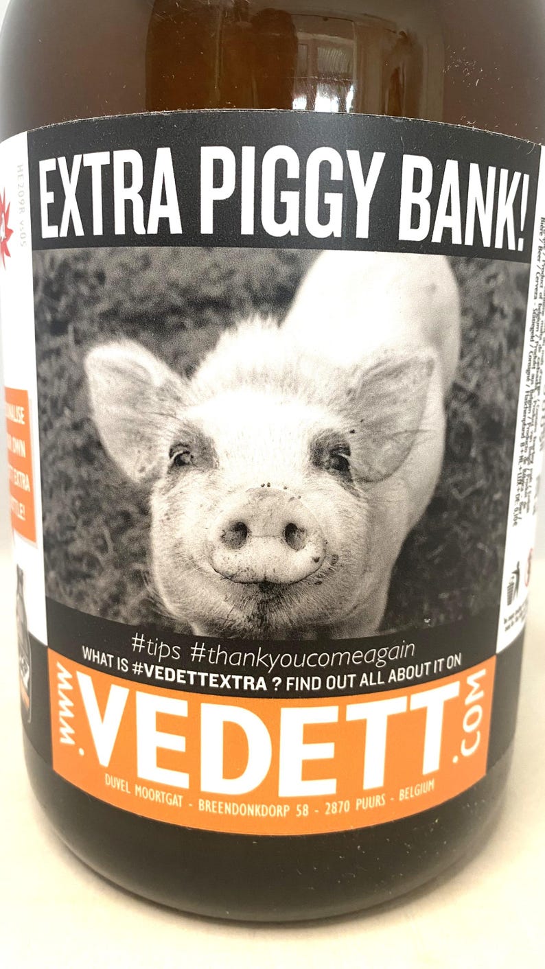 May include: A brown bottle with a black and orange label featuring a black and white image of a pig's face. The label reads "EXTRA PIGGY BANK!" and includes the text "#tips #thankyoucomeagain".