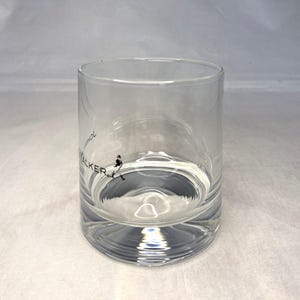May include: A clear, cylindrical glass with a thick base. The glass features the text "John Walker" in black, along with a silhouette of a man walking. The glass is partially filled with a clear liquid.