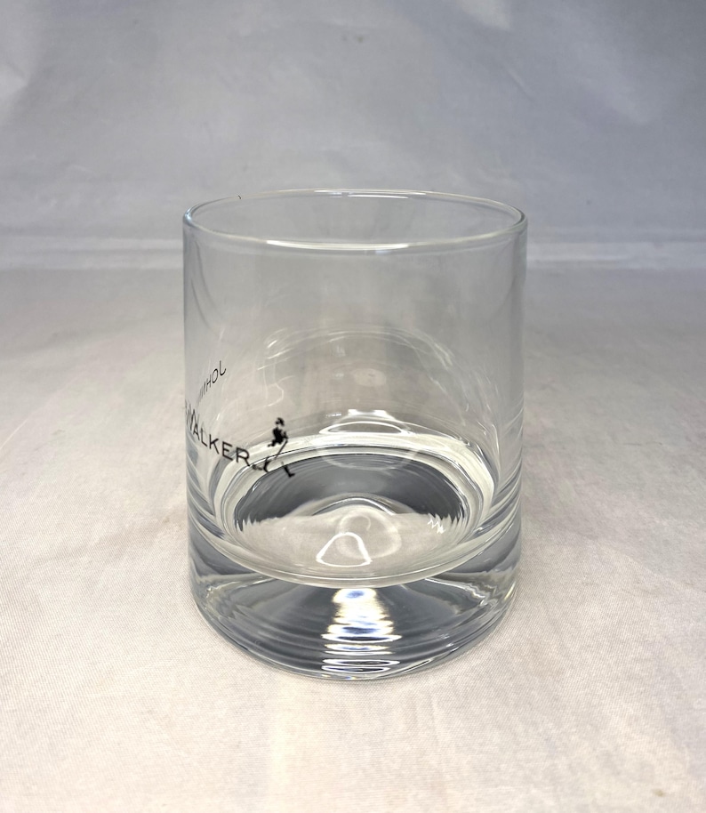 May include: A clear, cylindrical glass with a thick base. The glass features the text "John Walker" in black, along with a silhouette of a man walking. The glass is partially filled with a clear liquid.
