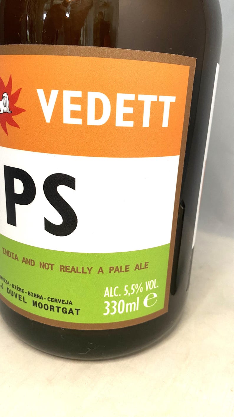 May include: A close-up of a beer bottle with a label featuring the word "VEDETT" in white on an orange background. The label also includes the text "PS" in black, and "INDIA AND NOT REALLY A PALE ALE". The bottle contains 330ml.