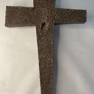 May include: A dark gray cross made of rough, textured material. The cross has a unique, asymmetrical design with a pointed top.