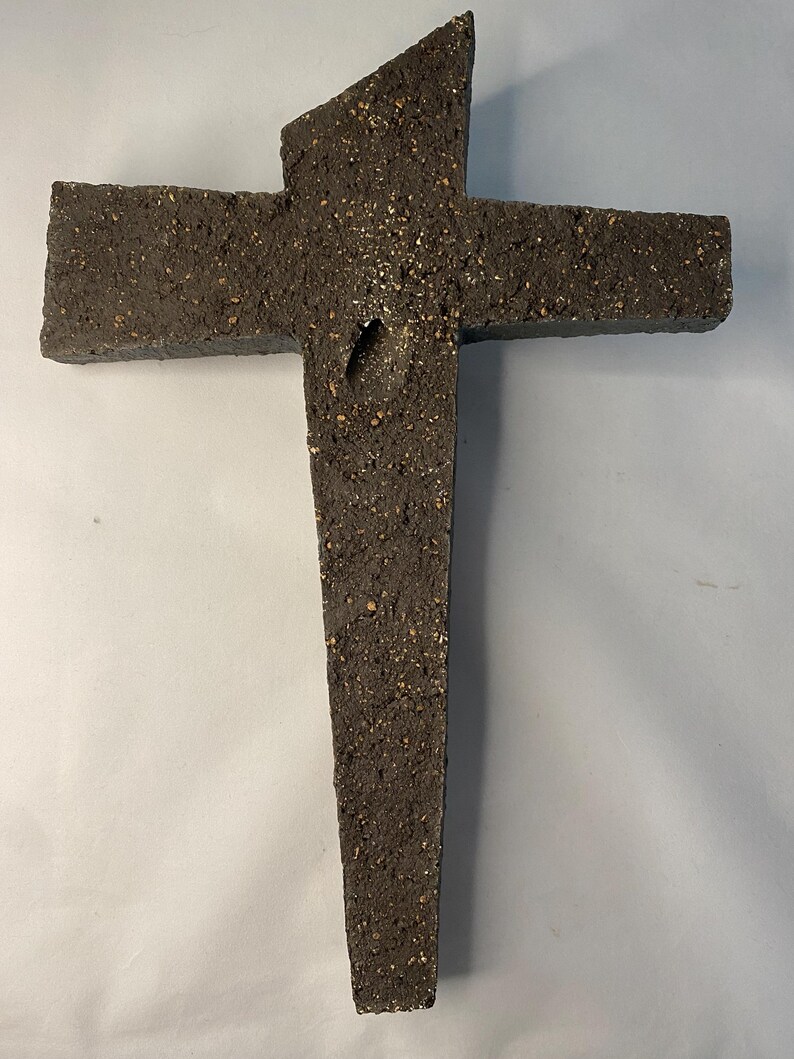May include: A dark gray cross made of rough, textured material. The cross has a unique, asymmetrical design with a pointed top.