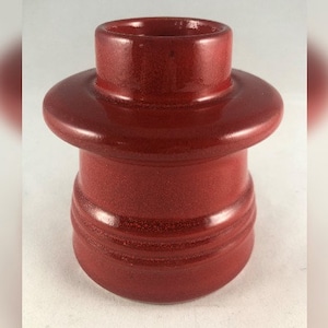 Könnte beinhalten: Ein roter Kerzenhalter aus Keramik mit einer breiten Basis und einer kleineren oberen Öffnung. Der Halter hat eine strukturierte Oberfläche und eine glänzende Oberfläche.