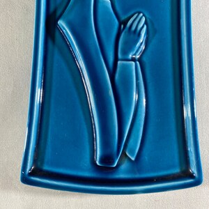 May include: A blue ceramic wall plaque with a raised image of a woman in prayer. The woman is wearing a long robe and has her hands clasped in front of her.
