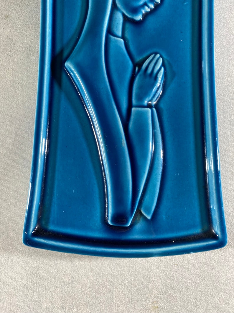 May include: A blue ceramic wall plaque with a raised image of a woman in prayer. The woman is wearing a long robe and has her hands clasped in front of her.