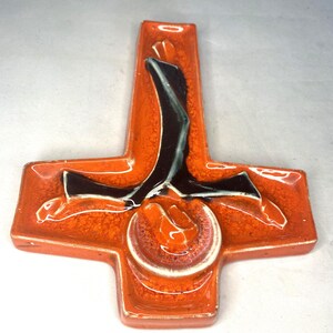 May include: Orange ceramic wall tile with a black and white design of a person in a cross shape.