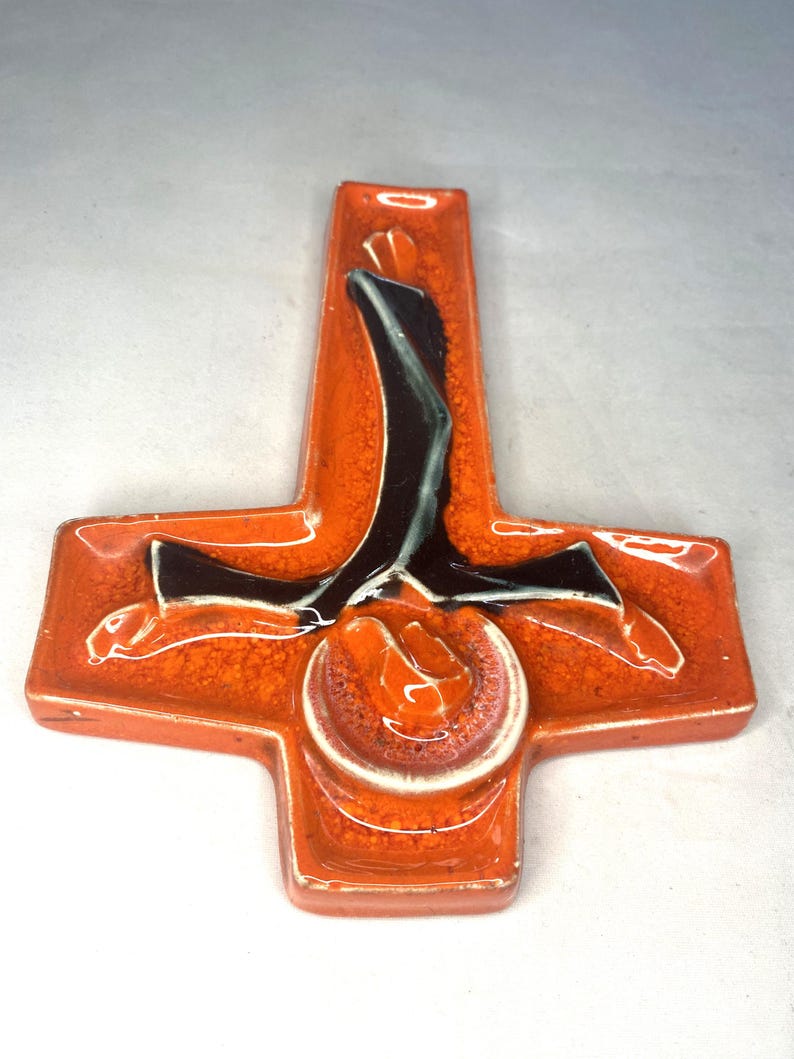 May include: Orange ceramic wall tile with a black and white design of a person in a cross shape.