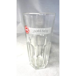 Può includere: Un bicchiere trasparente e sfaccettato con la scritta "JAMESON" stampata in bianco. Il bicchiere ha un logo rosso e bianco sul lato. Il bicchiere è vuoto e poggia su una superficie bianca.