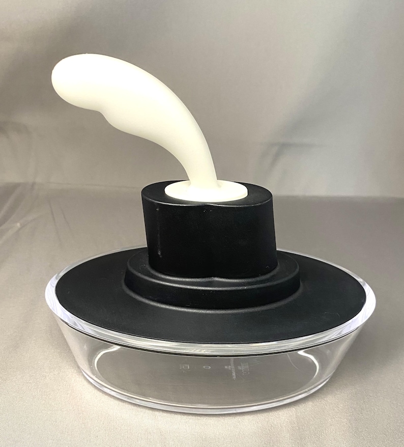 May include: A clear glass container with a black lid and a white, curved handle. The handle is attached to a black cylinder on top of the lid. The container has measurement markings on the side.