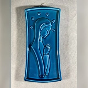 May include: A blue ceramic wall plaque with a raised image of a woman in prayer. The plaque is decorated with stars.