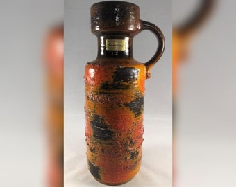 Może przedstawiać: Brązowy i pomarańczowy ceramiczny wazon z czarno-pomarańczowym geometrycznym wzorem. Wazon ma uchwyt i etykietę z napisem "Carstens Keramik".
