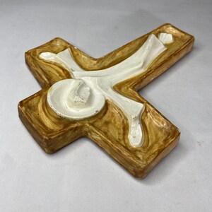 May include: A decorative cross with a textured, light brown base and a white, stylized figure. The cross has a three-dimensional design with rounded edges and a glossy finish, suitable for religious decor.