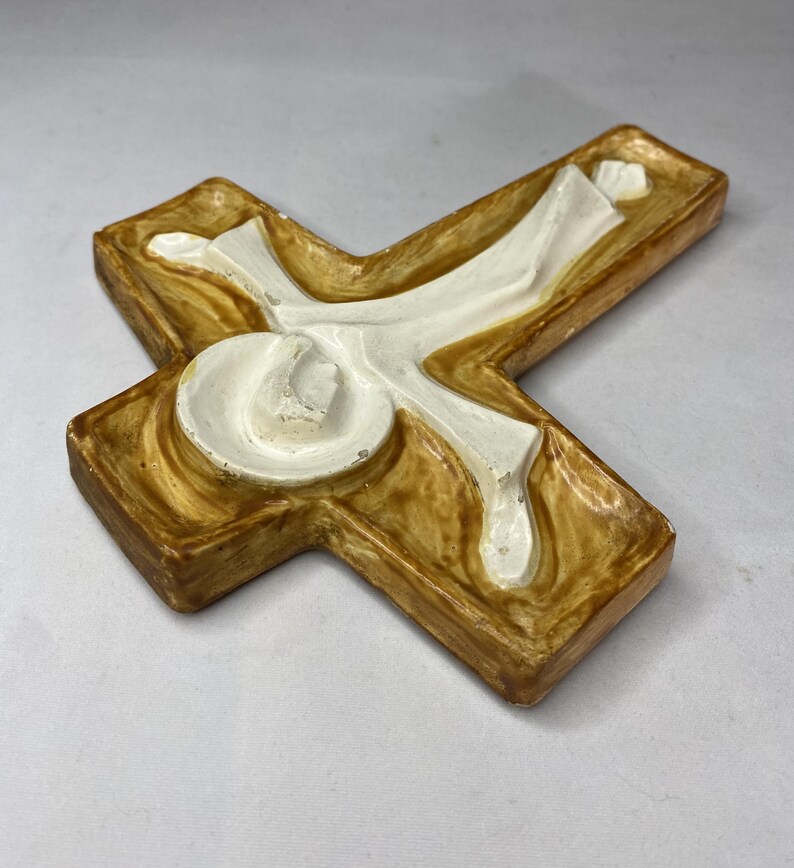 May include: A decorative cross with a textured, light brown base and a white, stylized figure. The cross has a three-dimensional design with rounded edges and a glossy finish, suitable for religious decor.