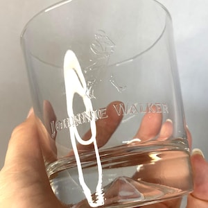 Puede incluir: Vaso bajo transparente con el texto grabado "JOHNNIE WALKER" y un logotipo de un hombre caminando. El vaso est&aacute; parcialmente lleno de un l&iacute;quido transparente. El vaso se sostiene en una mano.
