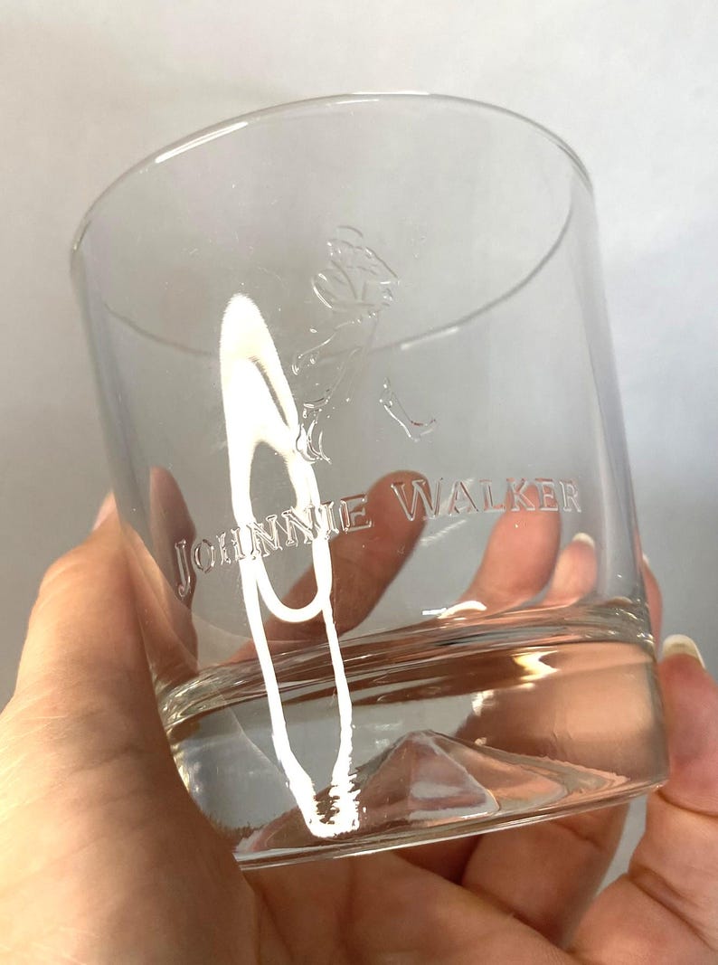 Puede incluir: Vaso bajo transparente con el texto grabado "JOHNNIE WALKER" y un logotipo de un hombre caminando. El vaso est&aacute; parcialmente lleno de un l&iacute;quido transparente. El vaso se sostiene en una mano.