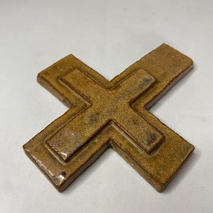 May include: A brown ceramic cross with a raised design. The cross is made of a textured clay and has a rustic look.