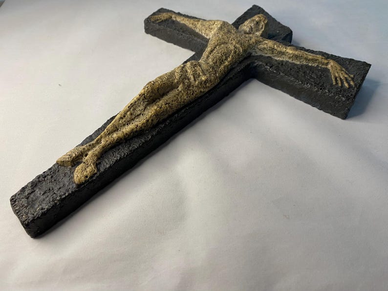 May include: A black ceramic cross with a beige sculpture of a person on it.