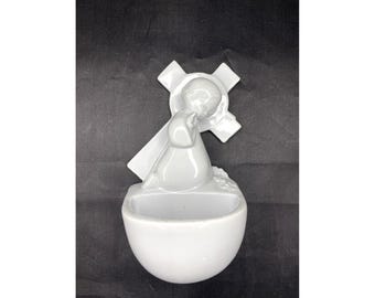May include: White ceramic wall-mounted holy water font with a figure of a person kneeling in prayer with a cross behind them.