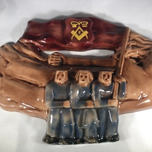 Pode incluir: Placa de parede de cerâmica representando três figuras com roupas azuis segurando uma faixa marrom com um símbolo maçônico dourado.