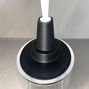 May include: A clear glass container with a black top and a white handle. The container has a wide, circular base and a cylindrical body. The black top has a tiered design. The white handle is long and slender.