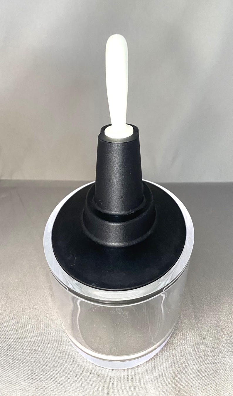 May include: A clear glass container with a black top and a white handle. The container has a wide, circular base and a cylindrical body. The black top has a tiered design. The white handle is long and slender.