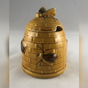 Könnte beinhalten: Ein Keramik-Honigtopf in Form eines Bienenstocks mit einer Biene oben drauf. Der Topf ist mit braunen und gelben Streifen verziert und hat eine kleine Öffnung zum Ausgießen von Honig.