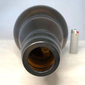 May include: A dark brown glass bottle with a wide opening, viewed from the top. A silver lighter is next to the bottle for scale. The bottle has a rounded shape and a smooth surface.