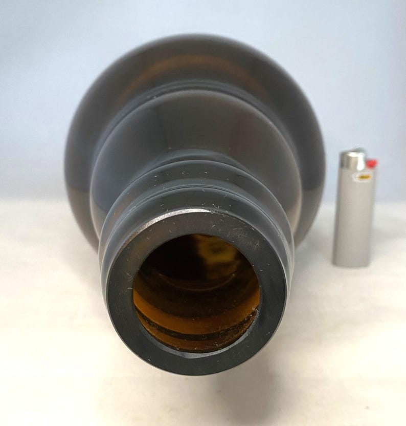 May include: A dark brown glass bottle with a wide opening, viewed from the top. A silver lighter is next to the bottle for scale. The bottle has a rounded shape and a smooth surface.