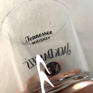 May include: A clear whiskey glass featuring the text "Tennessee Whiskey" and "Jack Daniel's" in black. The glass has a curved design and is held in a hand.