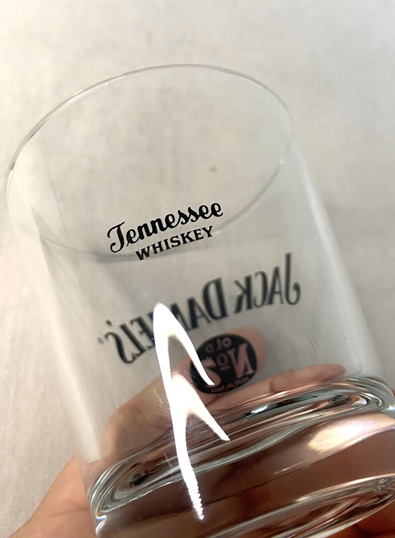 May include: A clear whiskey glass featuring the text "Tennessee Whiskey" and "Jack Daniel's" in black. The glass has a curved design and is held in a hand.