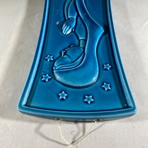 May include: A blue ceramic wall hanging with a raised image of a woman in a robe with stars around her. The wall hanging has two holes at the top for hanging.