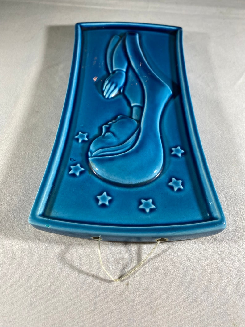 May include: A blue ceramic wall hanging with a raised image of a woman in a robe with stars around her. The wall hanging has two holes at the top for hanging.