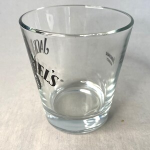May include: A clear shot glass with the text "DANIEL'S" in black. The glass has a slightly tapered design and a thick base. The shot glass is empty and appears to be made of glass.
